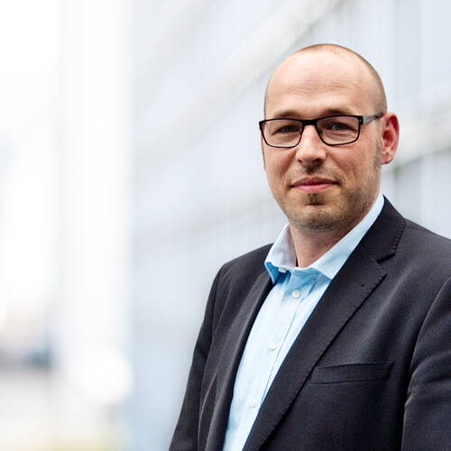 Glatzköpfiger Mann mit Brille im dunklen Anzug und hellblauem Hemd steht vor einem modernen Bürogebäude und blickt freundlich in die Kamera.