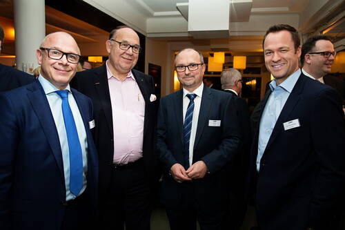 Vier lächelnde Geschäftsmänner in Anzügen stehen eng beisammen bei einer Abendveranstaltung in einem modern beleuchteten Konferenzraum.