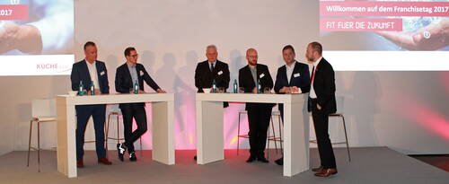 Sechs Männer im Anzug diskutieren auf einer Bühne beim Franchisetag 2017 an zwei Stehtischen vor einer Leinwand mit der Aufschrift „Willkommen auf dem Franchisetag 2017 – Fit für die Zukunft“.