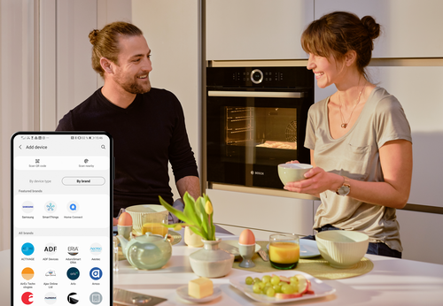 Paar bereitet gemeinsam Frühstück in moderner Küche zu, während ein vernetzter Bosch-Backofen läuft und ein Smartphone im Vordergrund die Einrichtung eines Smart-Home-Geräts zeigt.