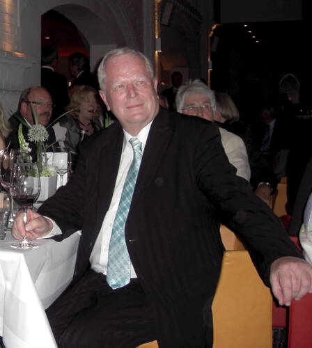 Älterer Mann im Anzug mit hellgrüner Krawatte sitzt entspannt an einem festlich gedeckten Tisch in einem gedämpft beleuchteten Saal und schaut lächelnd zur Seite.