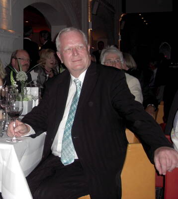 Älterer Mann im Anzug mit hellgrüner Krawatte sitzt entspannt an einem festlich gedeckten Tisch in einem gedämpft beleuchteten Saal und schaut lächelnd zur Seite.
