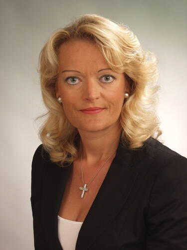 Blondgelockte Frau im schwarzen Blazer mit Kreuzkette blickt ernst in die Kamera vor neutralem Hintergrund