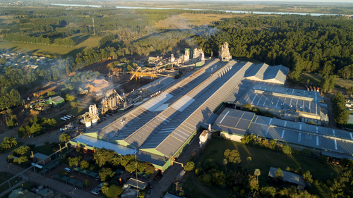 Großes Industrie- und Sägewerkgelände mit langen Hallen und rauchenden Schornsteinen, umgeben von dichtem Wald und einer ländlichen Siedlung im Hintergrund