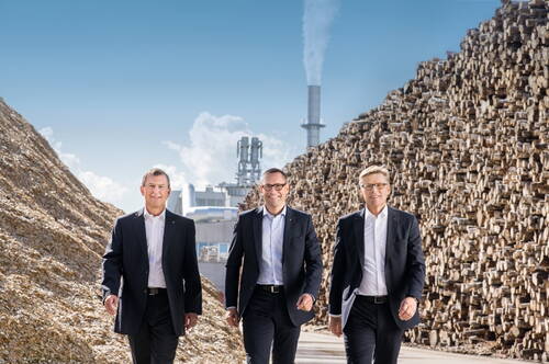 Drei Manager in Anzügen gehen zwischen riesigen Holzstapelbergen auf dem Gelände einer industriellen Produktionsanlage mit rauchendem Schornstein entlang