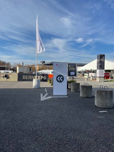 Wegweiser zur Swiss Interior Expo auf dem Außengelände der Bernexpo mit Flagge, Betonpollern und großem Messezelt unter blauem Himmel