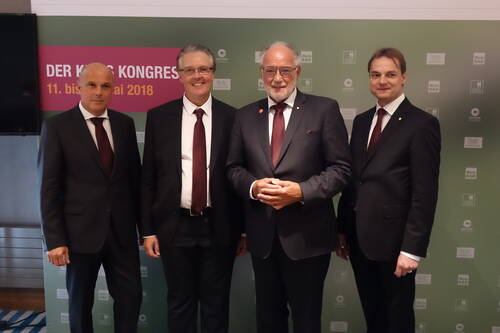 Vier Geschäftsleute in dunklen Anzügen mit bordeauxfarbenen Krawatten posieren lächelnd vor einer Kongresswand mit der Aufschrift „Der Kongress“ und dem Datum im Mai 2018.