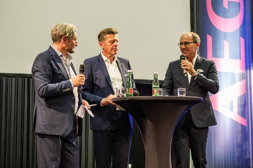 Drei elegant gekleidete Männer diskutieren auf einer Bühne an einem Stehtisch mit Wasserflaschen und Mikrofonen während einer Podiumsdiskussion.