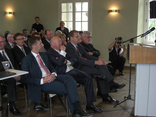 Geschäftsleute in Anzügen sitzen aufmerksam in Reihen und hören einem Redner an einem Podium mit Mikrofon in einem hellen Konferenzraum zu.