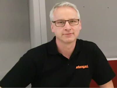 Mittelalterlicher Mann mit Brille in schwarzem Poloshirt mit orangefarbener Aufschrift „stengel“ sitzt vor graurotem Hintergrund und blickt neutral in die Kamera.
