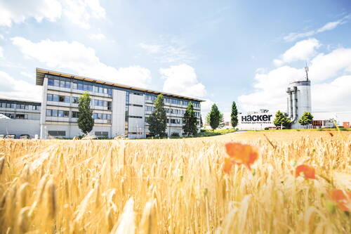 Modernes Firmengebäude und Produktionsanlage von Häcker Küchen hinter einem goldenen Getreidefeld mit Mohnblumen unter blauem Himmel