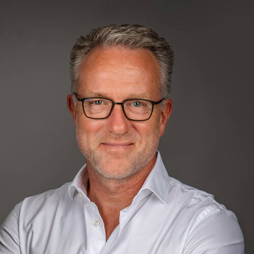 Mittelalterlicher Mann mit grauen Locken, Brille und leichtem Bart lächelt freundlich in weißem Hemd vor neutral grauem Hintergrund