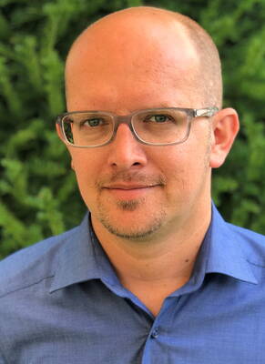 Glatzköpfiger Mann mit Brille und blauem Hemd blickt ruhig in die Kamera vor grünem, unscharfem Hintergrund.