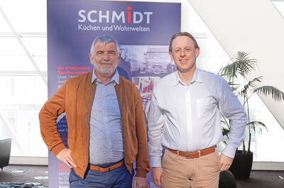 Zwei lächelnde Männer posieren in Geschäftskleidung vor einem Werbebanner des Küchen- und Wohnunternehmen Schmidt in einem hellen, modernen Innenraum.