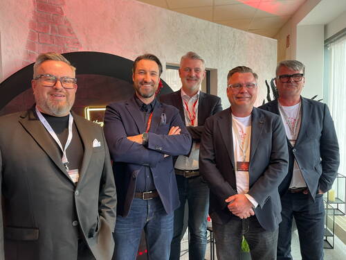 Fünf lächelnde Männer in Business-Kleidung posieren eng beieinander in einem modern gestalteten Konferenzraum mit Namensschildern um den Hals