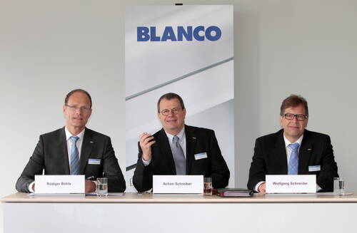 Drei Manager in Anzügen sitzen bei einer Pressekonferenz vor einem BLANCO‑Firmenschild an einem Tisch mit Namensschildern, Wasser und Unterlagen.