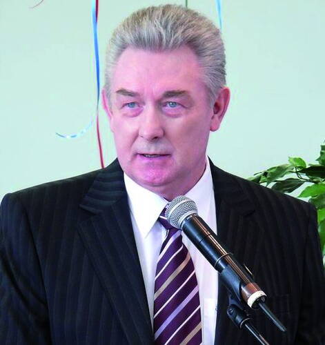 Älterer Mann im dunklen Nadelstreifenanzug spricht ernsthaft in ein Mikrofon vor hellem Hintergrund mit Dekoration und Pflanze.