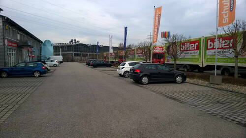Gewerbepark mit geparkten Autos und mehreren bunt beklebten Möbeltransportern entlang einer breiten Zufahrtsstraße an einem bewölkten Tag