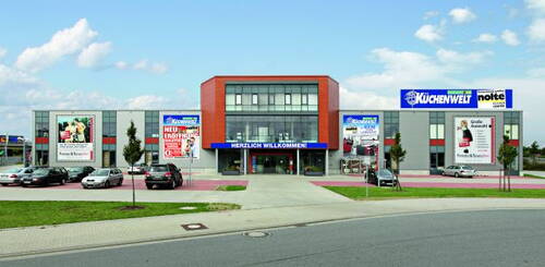 Modernes Küchenfachgeschäft mit großer Glasfassade, breitem Eingangsbereich mit „Herzlich willkommen“-Banner, Werbetafeln an den Seitenflügeln und mehreren parkenden Autos auf dem Parkplatz davor