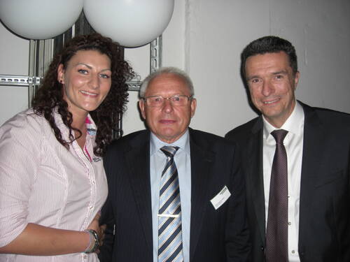 Drei elegant gekleidete Menschen stehen lächelnd nebeneinander bei einer Veranstaltung in Innenräumen, zwei Männer im Anzug und eine Frau im Businesshemd vor einer weißen Wand mit Ballons im Hintergrund.