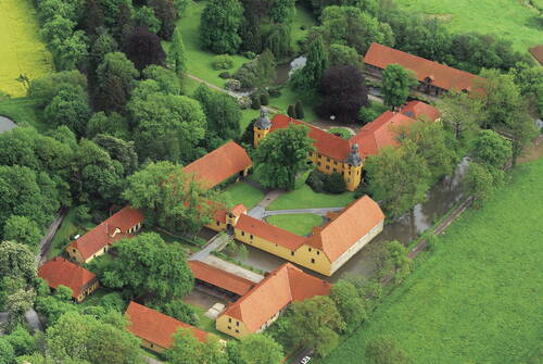 Historische Wasserburg mit gelben Gebäuden und roten Dächern, von breiten Gräben und dichtem, leuchtend grünem Parkland umgeben, aus der Luft betrachtet