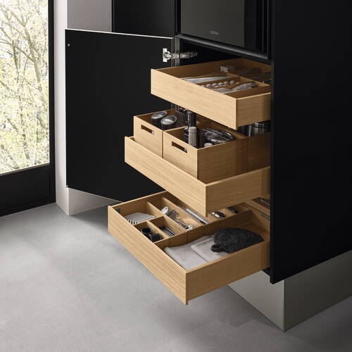 Mehrere geöffnete, ausziehbare Holzschubladen in einem modernen schwarzen Badschrank zeigen ordentlich sortierte Pflegeprodukte, Kosmetik und Handtücher.