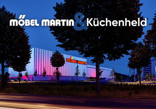 Leuchtend pink und blau illuminierte Möbel-Martin-Filiale bei Abenddämmerung hinter einer Baumreihe an einer vorbeiführenden Straße