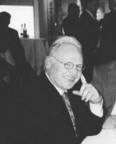 Älterer Mann im Anzug sitzt lächelnd an einem Tisch in einem eleganten, stimmungsvoll beleuchteten Raum und stützt den Kopf nachdenklich auf die Hand.