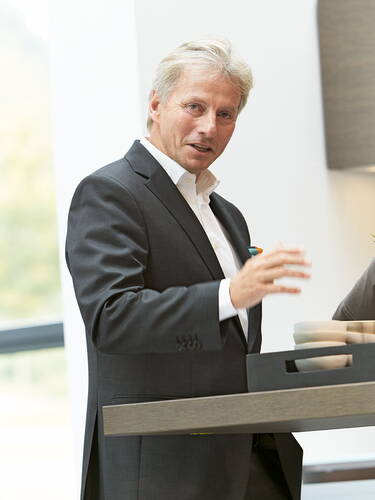 Älterer Mann im Anzug spricht gestikulierend an einem Stehtisch in einem hellen, modernen Raum