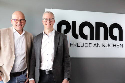 Zwei lächelnde Männer in Businesskleidung stehen vor einem Firmenlogo mit der Aufschrift „plana – Freude an Küchen“ in einem modernen Büro.