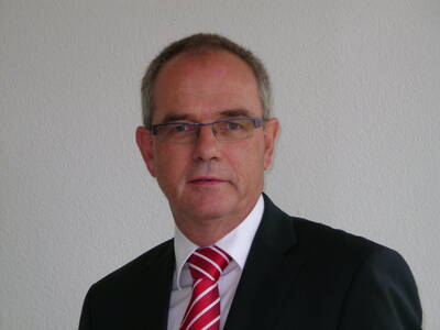Älterer Mann mit Brille im dunklen Anzug und rot-weiß gestreifter Krawatte steht vor einer hellen Wand und blickt ernst in die Kamera.
