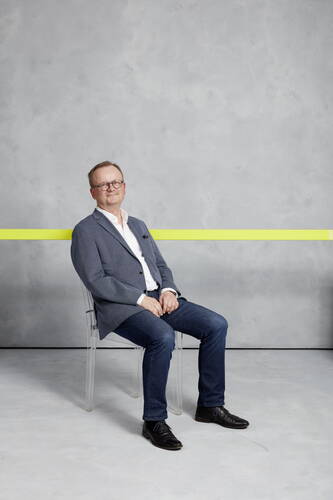 Mittelalterlicher Mann mit Brille sitzt entspannt auf einem transparenten Stuhl vor grauer Wand mit gelbem waagerechtem Streifen, trägt blaues Sakko, weiße Hemdbluse und Jeans.