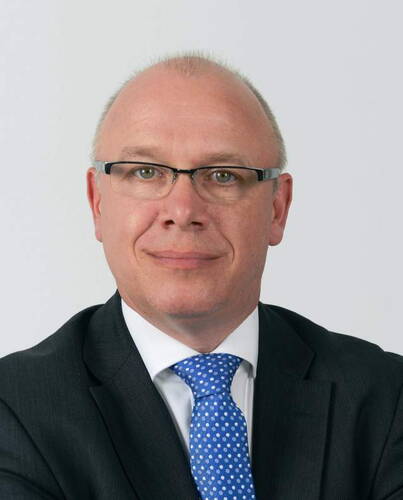 Glatzköpfiger Mann mit Brille im dunklen Anzug und blauer Krawatte blickt ernst in die Kamera vor neutralem Hintergrund.