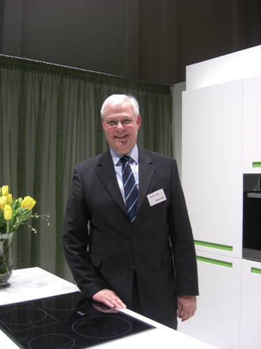 Älterer Mann im Anzug steht lächelnd neben einem modernen Induktionskochfeld in einer hellen Ausstellungsküche mit gelben Tulpen auf der Arbeitsplatte.