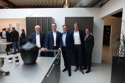 Fünf geschäftlich gekleidete Männer stehen lächelnd in einer modernen Ausstellungsküche vor einer Kücheninsel mit Induktionskochfeld und großer schwarzer Vase, während im Hintergrund Gäste miteinander ins Gespräch vertieft sind.