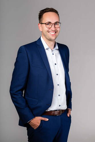 Lächelnder Mann mit Brille in blauem Anzug und weißem Hemd steht vor neutralem Hintergrund und hat eine Hand in der Hosentasche
