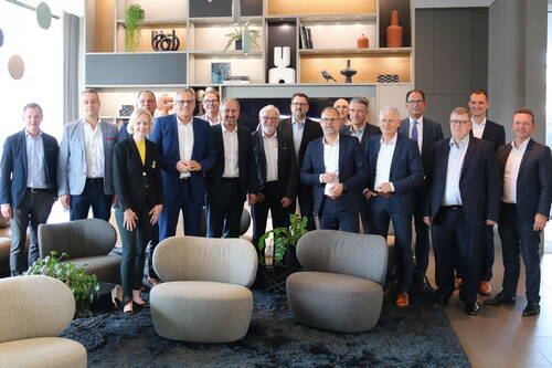 Gruppe von Geschäftsleuten in Anzügen und Businesskleidung posiert lächelnd in einem modern eingerichteten Besprechungsraum mit Designer-Sitzmöbeln und dekorativen Regalen im Hintergrund