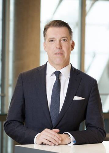 Mittelalter Mann im dunklen Anzug steht an einem Tisch und blickt ruhig in die Kamera, im Hintergrund ein modernes Bürogebäude mit großen Glasfronten