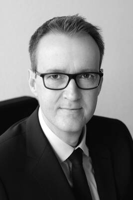 Porträt eines Mannes mittleren Alters mit Kurzhaarfrisur und Brille im Anzug, der in schwarz-weiß freundlich in die Kamera blickt