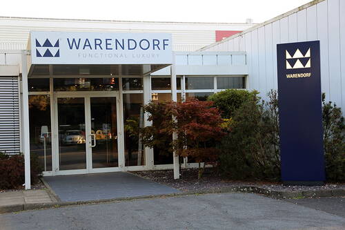 Eingang des Warendorf-Showrooms mit Glasfassade, beleuchtetem Firmenlogo und von Ziersträuchern umgebenem Vorplatz