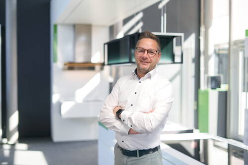 Mann mit Brille und weißem Hemd steht lächelnd mit verschränkten Armen in einem modernen, lichtdurchfluteten Büro oder Showroom.