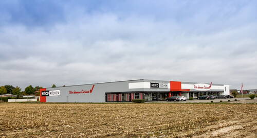 Modernes HEICO-Küchenfachgeschäft mit großer Glasfront und Parkplatz vor einem abgeernteten Feld unter bewölktem Himmel