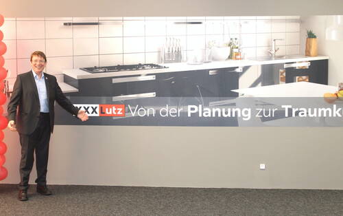 Lächelnder Mann im Anzug präsentiert vor einer Wand mit XXXLutz-Werbung und großem Küchenmotiv den Slogan „Von der Planung zur Traumküche“.