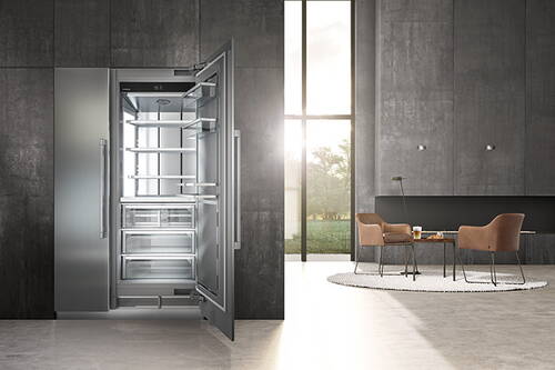 Offener, leerer Edelstahl-Kühlschrank in einer modernen, minimalistisch eingerichteten Wohnküche mit bodentiefem Fenster und Sitzbereich im Hintergrund