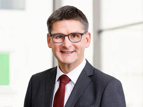Lächelnder Geschäftsmann mit Brille im dunklen Anzug und roter Krawatte steht vor einem hellen, unscharfen Bürohintergrund