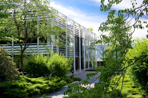 Modernes Bürogebäude mit gläserner Fassade und weißen Lamellen, umgeben von dichtem Grün und Bäumen an einem sonnigen Tag