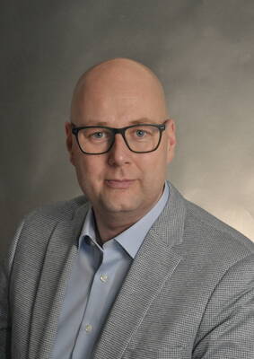 Glatzköpfiger Mann mit schwarzer Brille im grauen Sakko und hellblauem Hemd blickt frontal vor neutralem Hintergrund in die Kamera