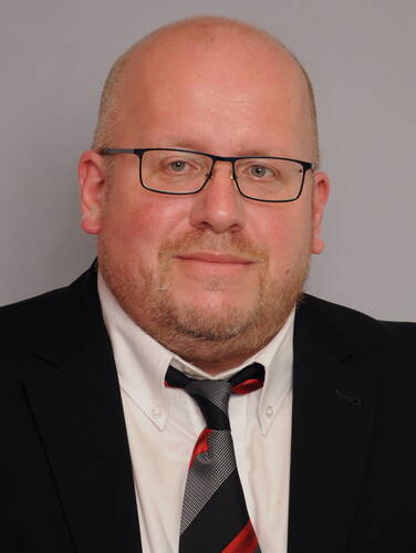 Glatzköpfiger Mann mit Brille im schwarzen Anzug und gestreifter rot‑schwarzer Krawatte vor neutral grauem Hintergrund