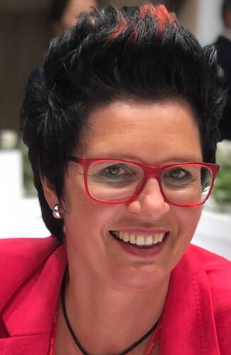 Lächelnde Frau mit kurzen schwarzen Haaren und roten Strähnen trägt rote Brille und roten Blazer in einer hellen Innenraumszene