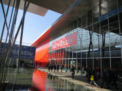 Besucher gehen an einem modernen Messegebäude mit roter Fassade und großem „Salone Bagno“-Schriftzug entlang, der sich in einem flachen Wasserbecken spiegelt